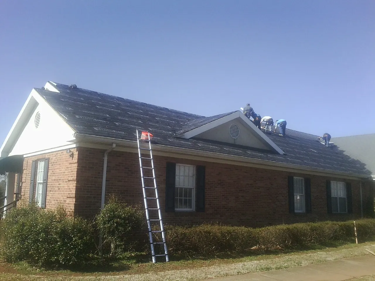 Crew tearing shingles off a brick ranch roof with a ladder set — Metal Roof Repair work by our crew in Goodrich